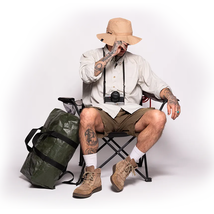 Man sitting on a chair with a bag and camera, wearing a hat. HERO 2024.